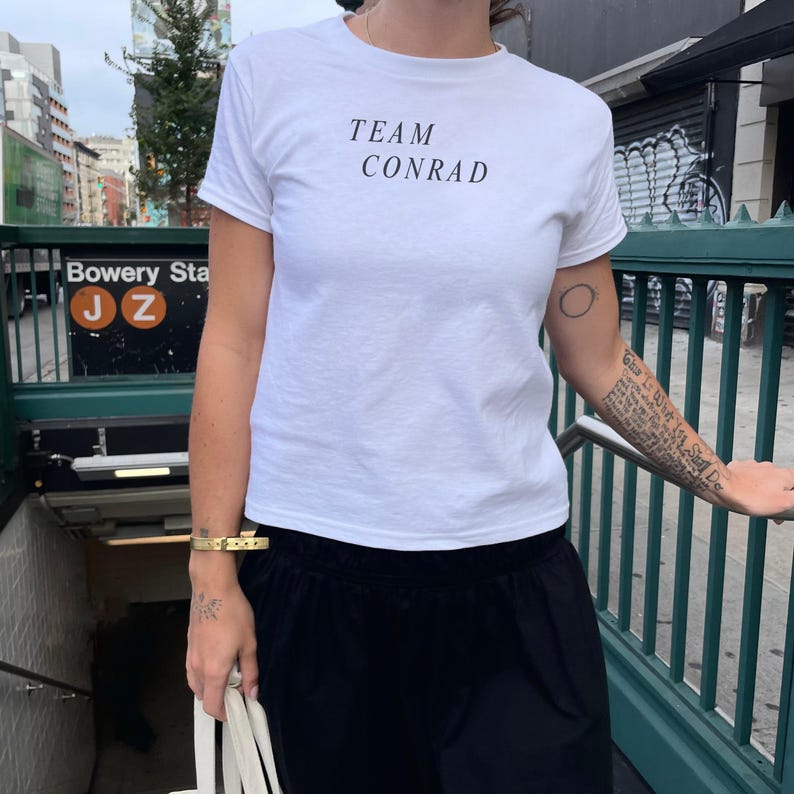 May include: A white t-shirt with the text "TEAM CONRAD" in black. The shirt is paired with black pants. The image is taken in front of a subway entrance.