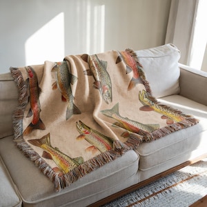 Może przedstawiać: Wełniana narzuta tkana z beżowym tłem i wzorem brązowych pstrąg&oacute;w. Narzuta ma frędzlowany brzeg.