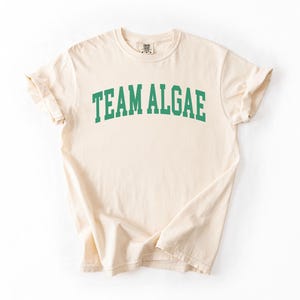 May include: A cream-colored short-sleeve t-shirt featuring the text "TEAM ALGAE" printed in a bold, arched, green collegiate-style font across the chest. The garment has a crew neckline and a standard fit with visible fabric texture.
