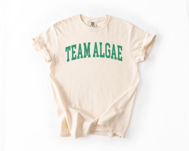 May include: A cream-colored short-sleeve t-shirt featuring the text "TEAM ALGAE" printed in a bold, arched, green collegiate-style font across the chest. The garment has a crew neckline and a standard fit with visible fabric texture.