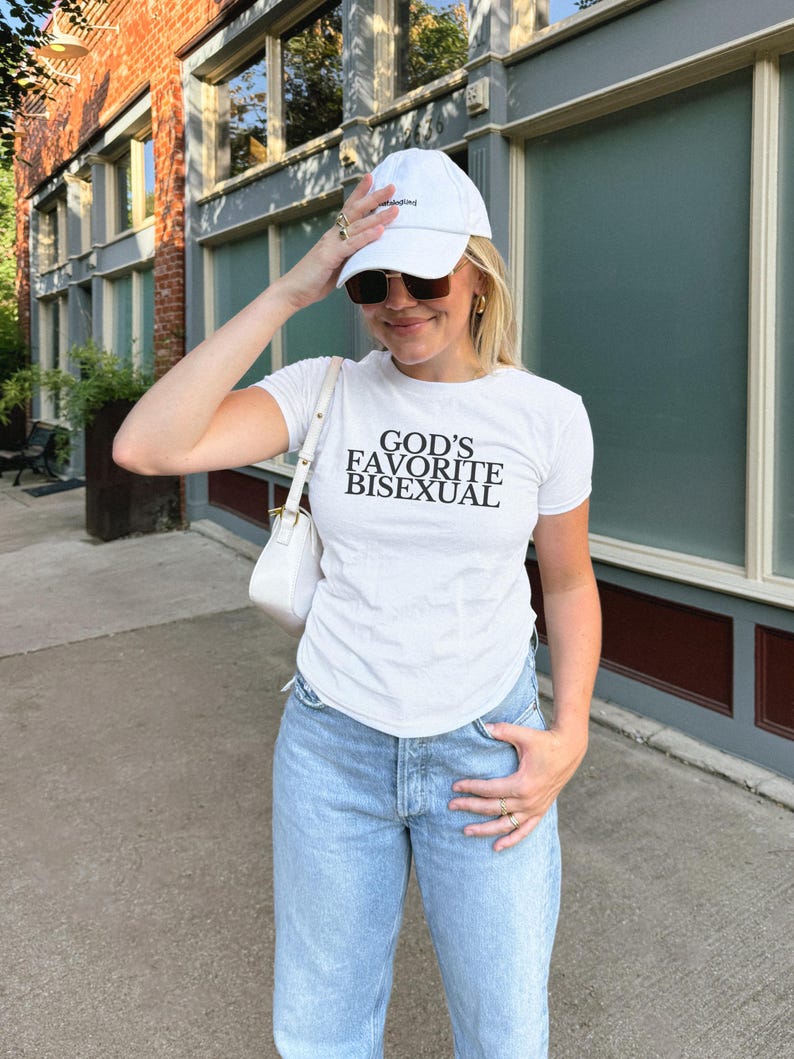 Op de afbeelding: Een persoon draagt een wit T-shirt met de tekst "GOD'S FAVORITE BISEXUAL" erop gedrukt. Ze dragen ook een witte baseballpet, blauwe jeans en een zonnebril.