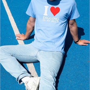 May include: Light blue t-shirt with the text "I ❤️ DIGGING IN MY BUTT" in white and red. The person is wearing light blue jeans, white sneakers, and a light blue cap. The background is a blue surface with a white stripe.