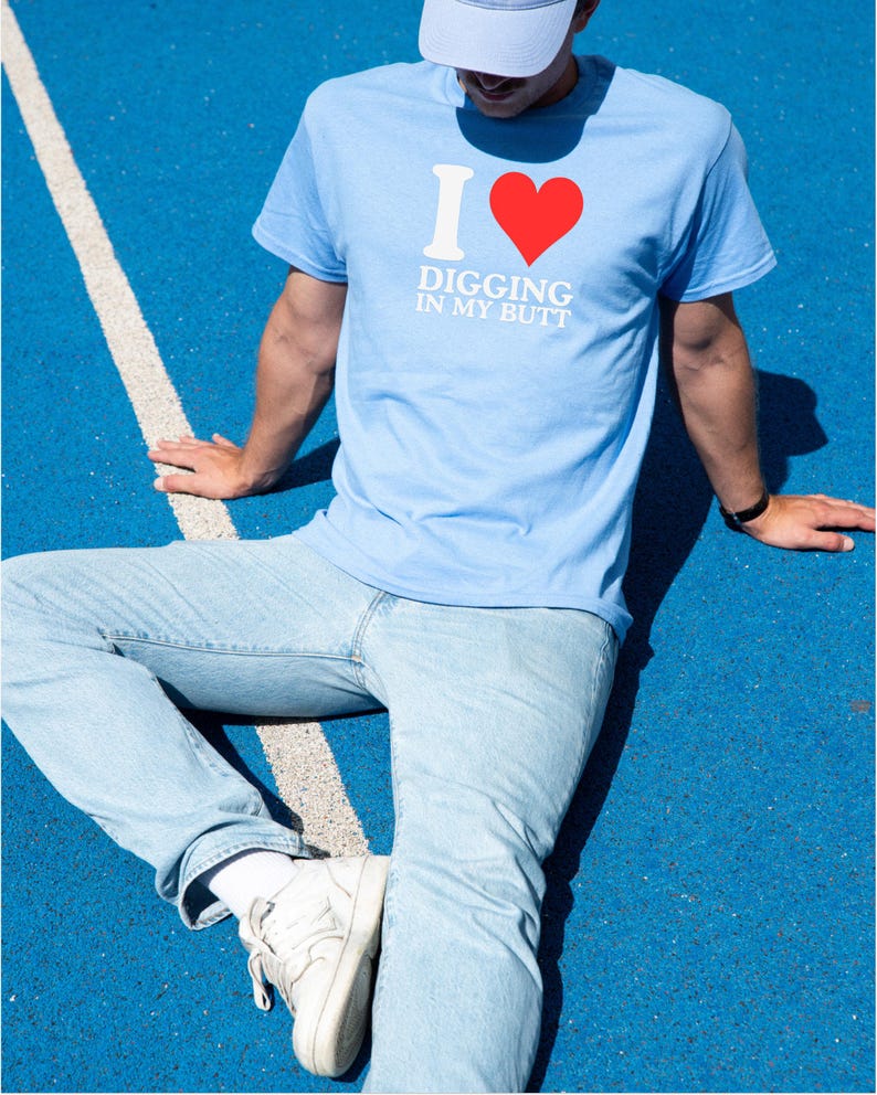 May include: Light blue t-shirt with the text "I ❤️ DIGGING IN MY BUTT" in white and red. The person is wearing light blue jeans, white sneakers, and a light blue cap. The background is a blue surface with a white stripe.