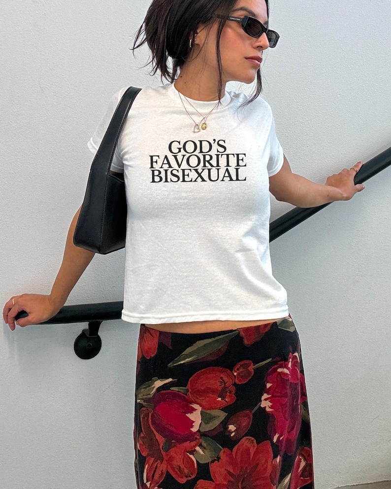 Op de afbeelding: Een wit T-shirt met de tekst "GOD'S FAVORITE BISEXUAL" erop gedrukt. De persoon die het shirt draagt, draagt ook een zwarte bloemenrok en een zonnebril.