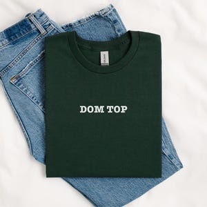 May include: A dark green t-shirt with the text "DOM TOP" printed in white on the front. The t-shirt is folded and placed on top of a pair of blue jeans.