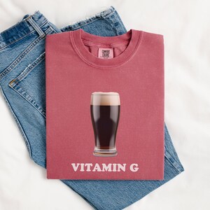 Pu&ograve; includere: Una maglietta rossa con un bicchiere di birra scura e la scritta "VITAMIN G" stampata su di essa.