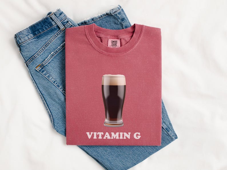 Pu&ograve; includere: Una maglietta rossa con un bicchiere di birra scura e la scritta "VITAMIN G" stampata su di essa.