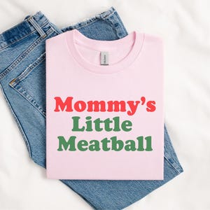 May include: A pale pink t-shirt featuring the text "Mommy's Little Meatball" in red and green. The shirt is folded and placed on top of a pair of blue denim jeans, against a white backdrop.