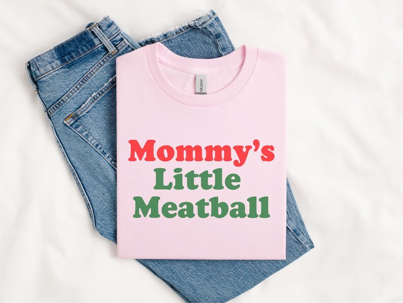 May include: A pale pink t-shirt featuring the text "Mommy's Little Meatball" in red and green. The shirt is folded and placed on top of a pair of blue denim jeans, against a white backdrop.