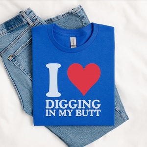 May include: A blue t-shirt with the text "I ❤️ DIGGING IN MY BUTT" in white and red. The shirt is folded and placed on top of a pair of blue jeans.