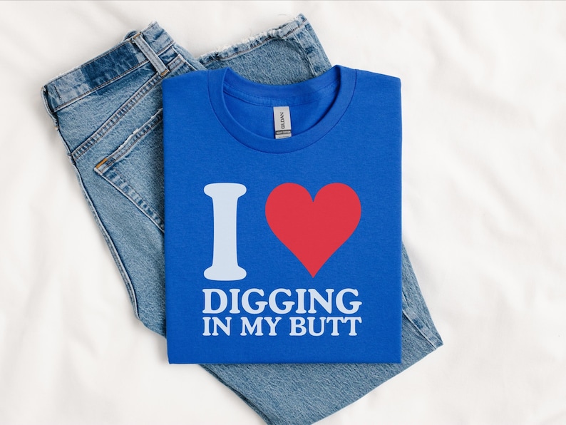May include: A blue t-shirt with the text "I ❤️ DIGGING IN MY BUTT" in white and red. The shirt is folded and placed on top of a pair of blue jeans.