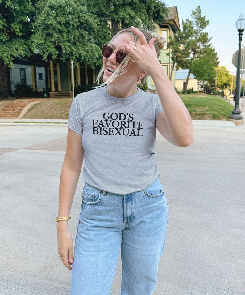 Op de afbeelding: Een vrouw draagt een grijs T-shirt met de tekst "God's Favorite Bisexual" erop gedrukt. Ze draagt een blauwe jeans en een zonnebril.