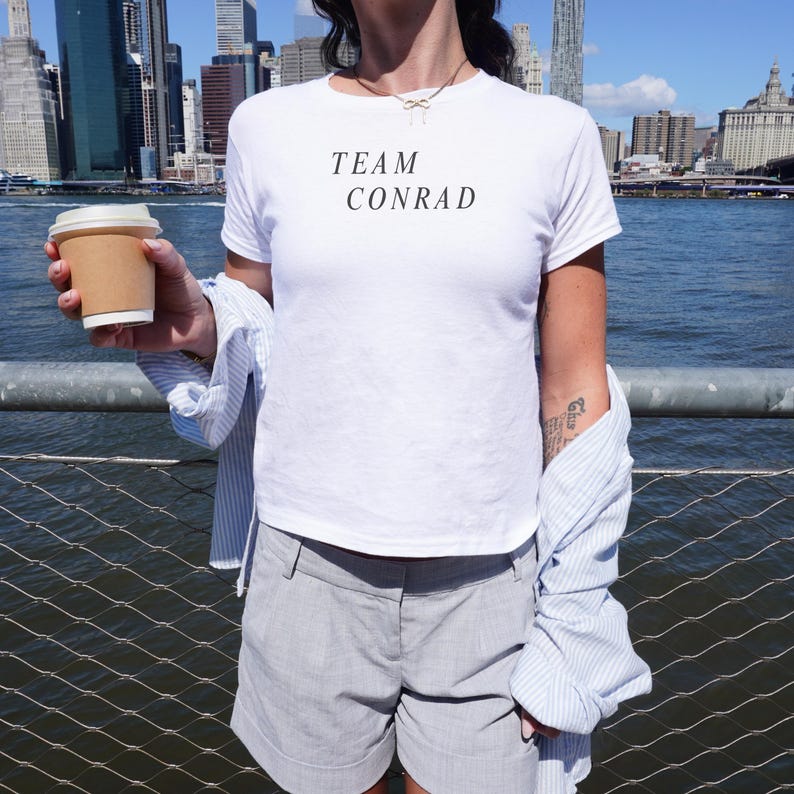 May include: White t-shirt with the text "TEAM CONRAD" in black. The person is wearing gray shorts and a light blue and white striped shirt. A disposable coffee cup is held in the hand.