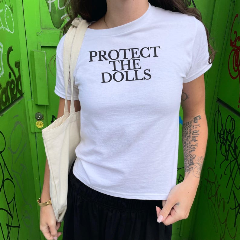 Puede incluir: Camiseta blanca de cuello redondo con el texto negro "PROTECT THE DOLLS". La camiseta se combina con pantalones negros y un bolso tote beige. El fondo es una puerta verde con graffiti.