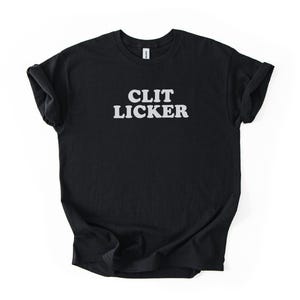 May include: Black t-shirt with the text "CLIT LICKER" in white, bold, capital letters. The shirt is made of a soft material and has rolled-up sleeves. The shirt is displayed on a white background.