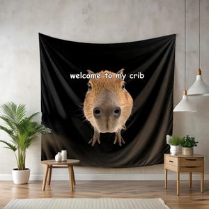 Puede incluir: Tapiz negro con una imagen de primer plano de la cara de un carpincho y el texto "welcome to my crib" en blanco. El carpincho tiene pelaje marrón y una nariz grande. El tapiz está colgado en una pared.