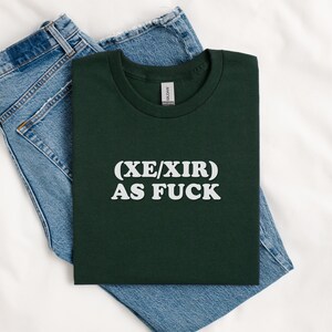 May include: A dark green t-shirt with the text "(XE/XIR) AS FUCK" printed in white.