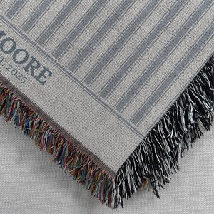 May include: A woven throw blanket with a striped pattern in shades of gray and white. The corner of the blanket is visible, showing a fringe of black and multicolored threads. The text "MOORE" and "EST. 2025" is printed on the blanket.
