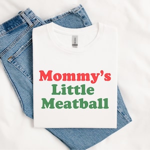 May include: White t-shirt with the text "Mommy's Little Meatball" in red and green. The shirt is laid on top of a pair of blue denim jeans. The shirt is a crew neck style.