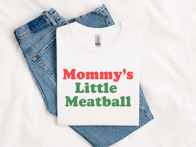 May include: White t-shirt with the text "Mommy's Little Meatball" in red and green. The shirt is laid on top of a pair of blue denim jeans. The shirt is a crew neck style.