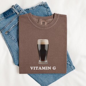 Pu&ograve; includere: Una maglietta marrone con una grafica di un bicchiere di birra scura e il testo "VITAMIN G". La maglietta &egrave; piegata e appoggiata su un paio di jeans blu.