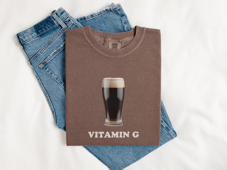 Pu&ograve; includere: Una maglietta marrone con una grafica di un bicchiere di birra scura e il testo "VITAMIN G". La maglietta &egrave; piegata e appoggiata su un paio di jeans blu.