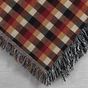 May include: A woven throw blanket with a plaid pattern in shades of red, brown, tan, black, and cream. The blanket has a fringe border with black and multicolored threads. The corner of the blanket is visible.