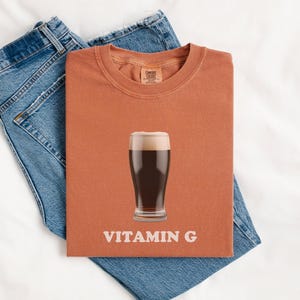 Pu&ograve; includere: Una maglietta color ruggine con una grafica di un bicchiere di birra scura e il testo "VITAMIN G".