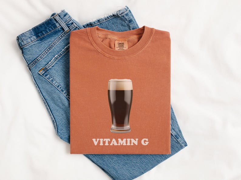 Pu&ograve; includere: Una maglietta color ruggine con una grafica di un bicchiere di birra scura e il testo "VITAMIN G".