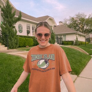 May include: A woman wearing a brown t-shirt with the text "Zombie Island Pepper Plantation & Inn" and a green chili pepper graphic. She is standing in front of a white building with a green lawn.