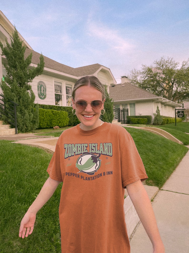 May include: A woman wearing a brown t-shirt with the text "Zombie Island Pepper Plantation & Inn" and a green chili pepper graphic. She is standing in front of a white building with a green lawn.