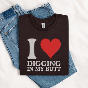May include: A dark brown t-shirt with the phrase "I ❤️ DIGGING IN MY BUTT" in white lettering and a red heart graphic. The shirt is folded on top of a pair of blue denim jeans, all resting on a white surface.