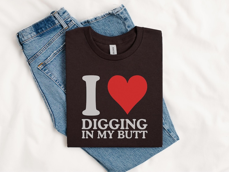 May include: A dark brown t-shirt with the phrase "I ❤️ DIGGING IN MY BUTT" in white lettering and a red heart graphic. The shirt is folded on top of a pair of blue denim jeans, all resting on a white surface.