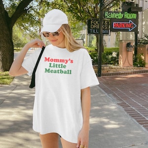 May include: White t-shirt with the text "Mommy's Little Meatball" in red and green. The shirt is worn by a person wearing a white cap and sunglasses. The background includes trees, a street, and a sign that says "Kennedy Room".