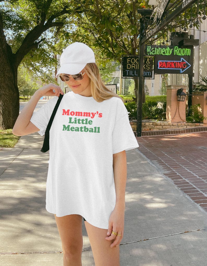 May include: White t-shirt with the text "Mommy's Little Meatball" in red and green. The shirt is worn by a person wearing a white cap and sunglasses. The background includes trees, a street, and a sign that says "Kennedy Room".