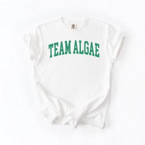 May include: A white short-sleeved t-shirt featuring the text "TEAM ALGAE" printed in a bold, arched green collegiate-style font across the chest. The garment has a crew neckline and a standard fit, showcasing a clean and simple design.