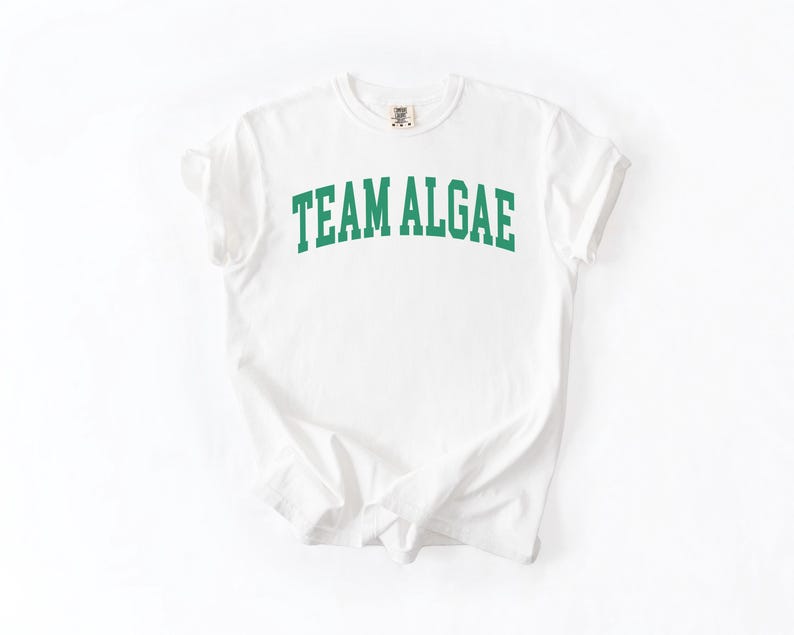 May include: A white short-sleeved t-shirt featuring the text "TEAM ALGAE" printed in a bold, arched green collegiate-style font across the chest. The garment has a crew neckline and a standard fit, showcasing a clean and simple design.
