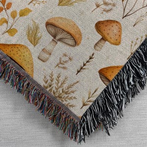 May include: A woven throw blanket with a cream background and a repeating pattern of brown mushrooms and stylised foliage. The blanket has a black fringe border.
