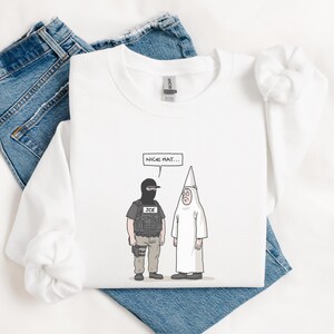 May include: White long-sleeve sweatshirt with a cartoon illustration. The graphic depicts two figures: one in a black mask and tactical vest with "ICE" on it, and another in a white robe and pointed hat. A speech bubble reads "NICE HAT..."