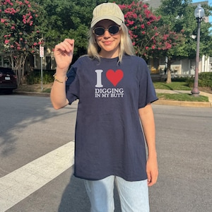 May include: Navy blue t-shirt with the text "I ❤️ DIGGING IN MY BUTT" in white and red. The shirt is worn with light-wash jeans, a tan cap, and sunglasses. The person is standing on a crosswalk.