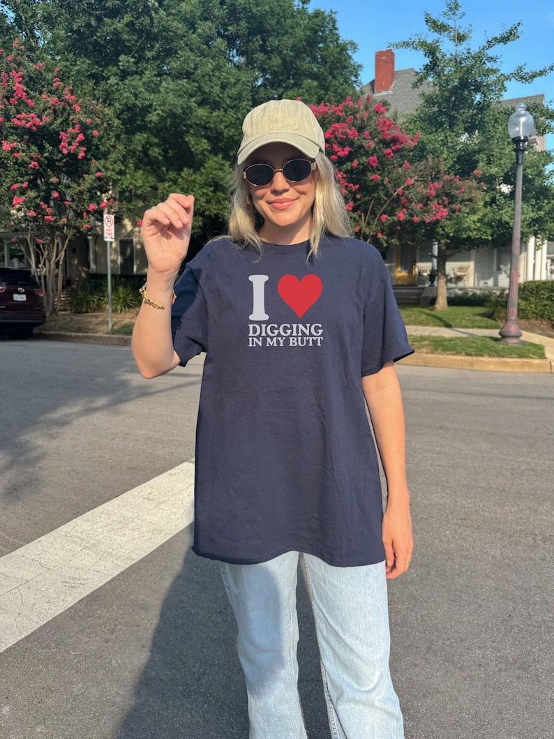 May include: Navy blue t-shirt with the text "I ❤️ DIGGING IN MY BUTT" in white and red. The shirt is worn with light-wash jeans, a tan cap, and sunglasses. The person is standing on a crosswalk.