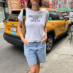 Puede incluir: Camiseta gris claro con el texto negro "PROTECT THE DOLLS". La camiseta se combina con pantalones cortos vaqueros azul claro. Un taxi amarillo es visible en el fondo.