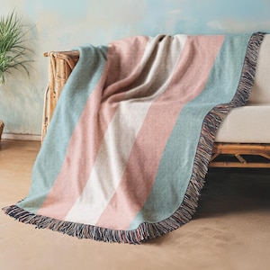 May include: A woven throw blanket featuring wide vertical stripes in muted shades of sage green, dusty rose, and cream. The blanket has a textured fabric finish and is finished with a decorative fringe along the edges, creating a soft appearance.