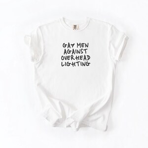 May include: White t-shirt with the text "Gay Men Against Overhead Lighting" printed in black.