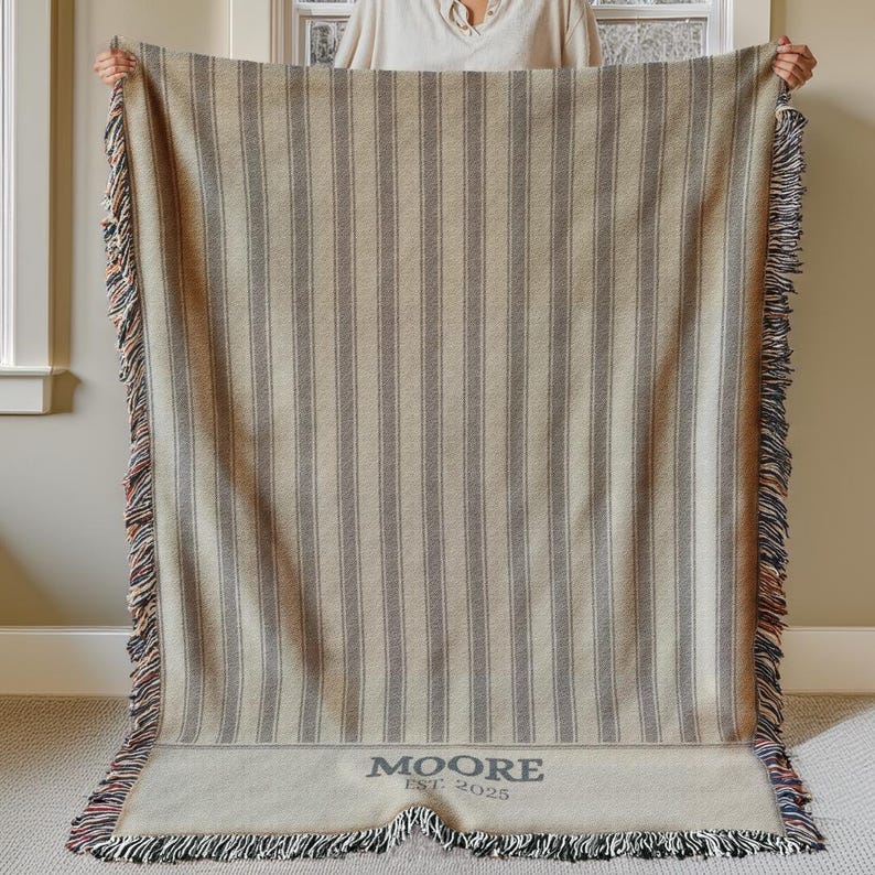 May include: A beige and gray striped throw blanket with a fringed edge. The blanket has vertical stripes and the text "MOORE EST. 2025" at the bottom. The fringe includes red, blue, and black threads.