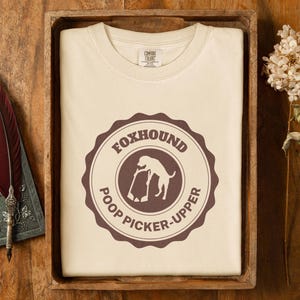 May include: A beige t-shirt with a brown circular graphic that says "Foxhound Poop Picker-Upper" and features a silhouette of a dog picking up poop.