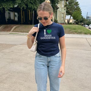 May include: A woman wearing a navy blue t-shirt with a white heart and the text "I &hearts; Irish Goodbyes". She is also wearing light blue jeans and sunglasses.