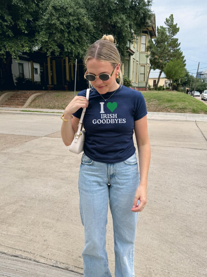 May include: A woman wearing a navy blue t-shirt with a white heart and the text "I &hearts; Irish Goodbyes". She is also wearing light blue jeans and sunglasses.