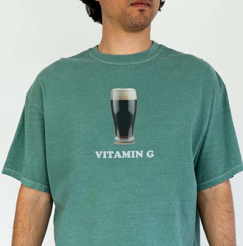 Pu&ograve; includere: Una maglietta verde con una grafica di un bicchiere di birra scura e il testo "VITAMIN G" stampato sul davanti.