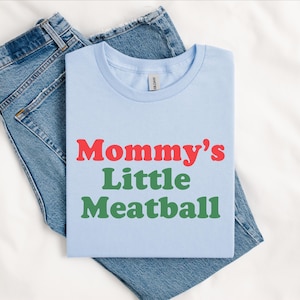 May include: Pale blue t-shirt featuring the text "Mommy's Little Meatball" in red and green. The t-shirt is folded and placed on a pair of blue denim jeans.